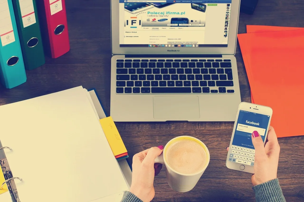 Person holding a coffee cup and iPhone displaying Facebook login beside a MacBook Air on a wooden desk with binders and papers.